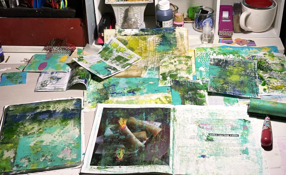 an art table with green and yellow monoprint art papers, a Gelli plate, a brayer, and other tools
